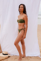 Model wearing Mila bandeau bikini too standing on a sandy beach with a white curtain in the background.